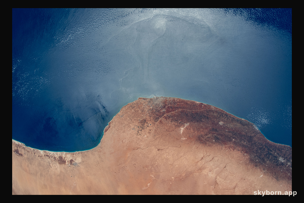 The sun's glint and the deep blue waters of the Mediterranean Sea stand in stark contrast to the desert coast of Libya