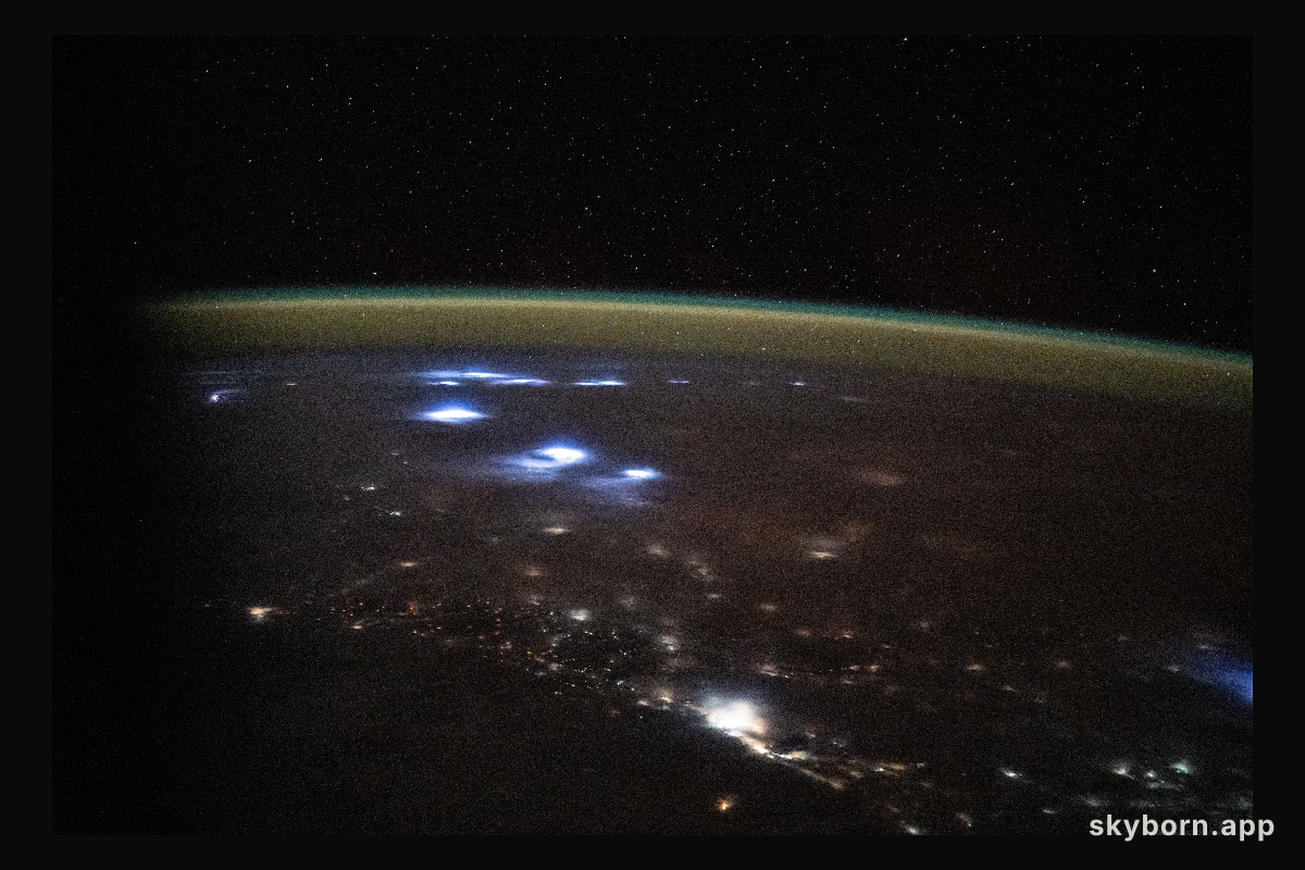 Warm, diffuse city lights and bright lightning storms illuminate the cloudy landscape